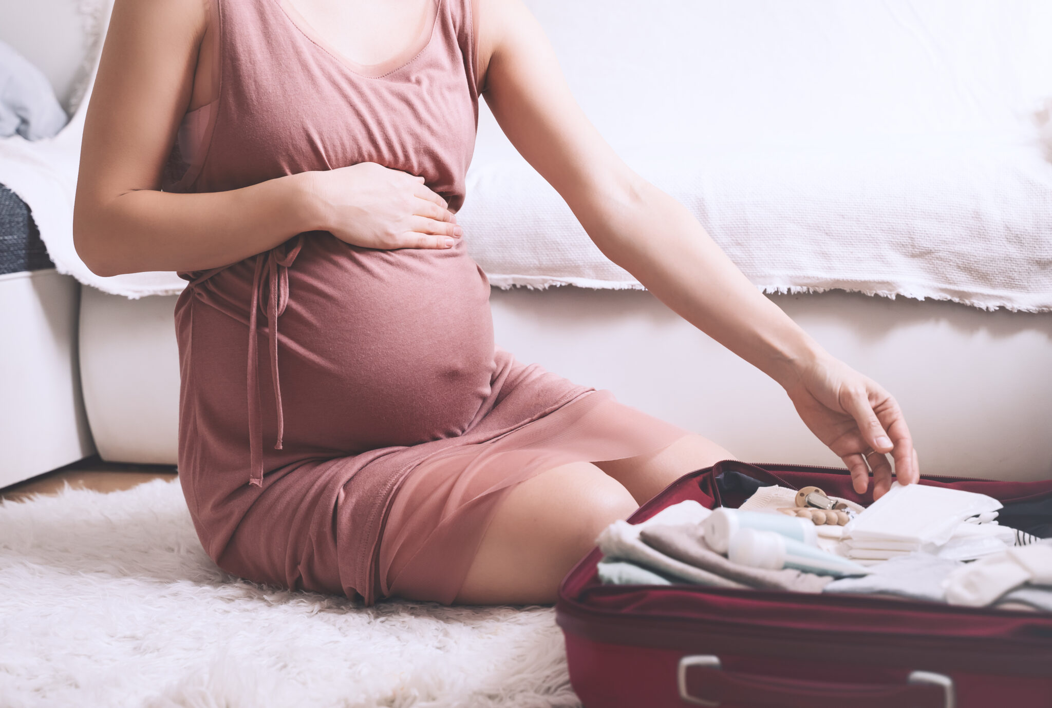 Close-up belly of pregnant woman in dress with travel bag of clothes and necessities. Сareful mother during pregnancy preparing and packing suitcase for maternity hospital getting ready for baby birth.